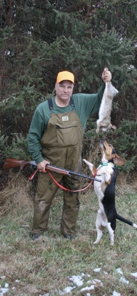 Hunter posing with a rifle, a rabbit, and a dog in a forested area.